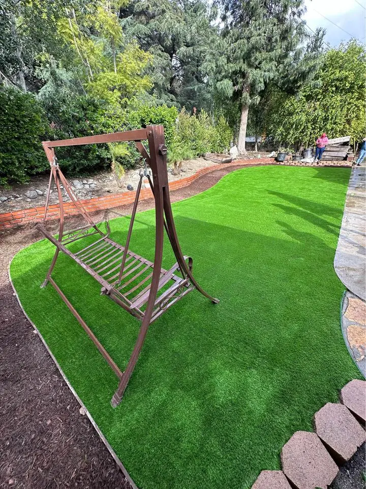A metal swing frame stands on bright artificial grass from expert turf solutions in a landscaped backyard with trees, bushes, a brick border, and a stone path. A person is visible in the background near the trees in Chandler AZ.