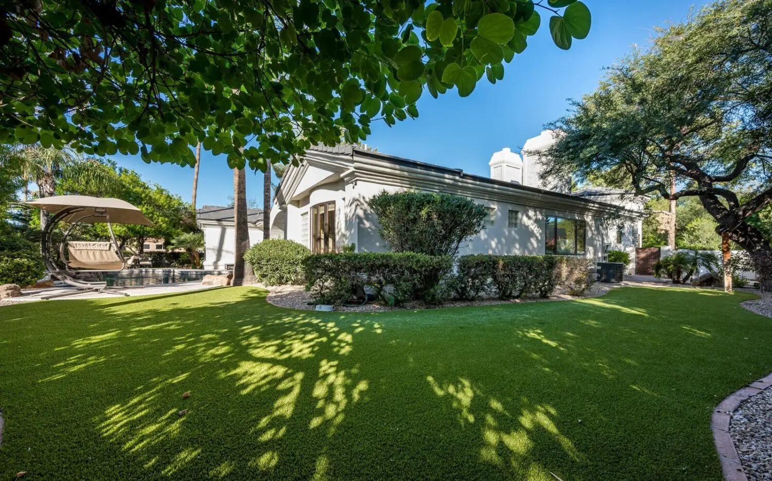 A landscaped backyard in Chandler AZ features green artificial grass, trimmed bushes, a large shade tree, and a hanging lounge chair by the pool—all thanks to expert turf solutions—under a clear blue sky.