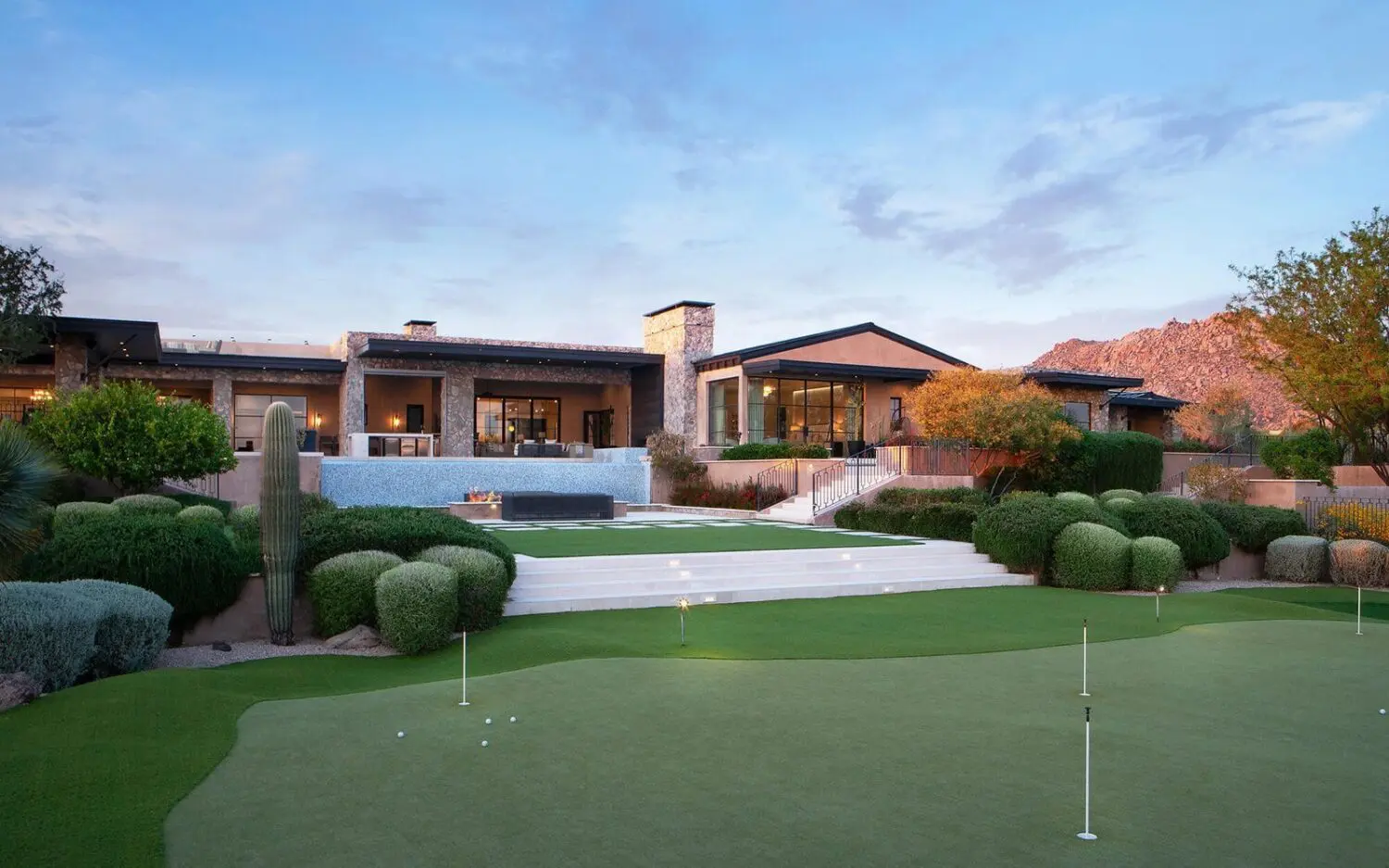 A modern luxury house with large windows and stone accents overlooks a manicured backyard featuring artificial grass putting green, trimmed bushes, stairs, and outdoor lounge areas at sunset in San Tan Valley.