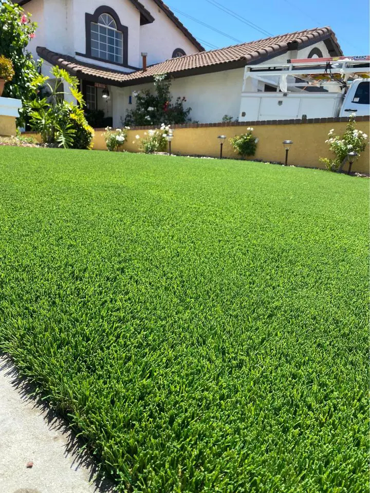 A neatly manicured artificial turf lawn in front of a house with white walls, brown roof tiles, flowering bushes, and a white truck parked in the driveway showcases quality artificial grass installation in Mesa AZ.