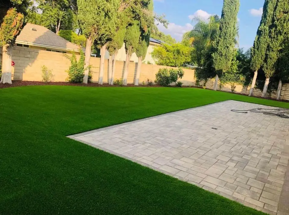 A backyard in Gilbert, AZ with a neatly mowed green lawn—ideal for those considering turf options or seeking an artificial grass installer—features a large paved patio, tall trees, and a stone wall under a partly cloudy sky.