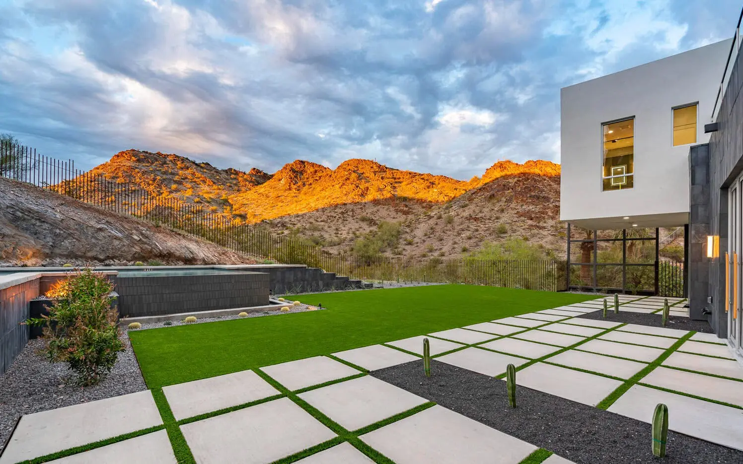 Modern house with large windows and a patio featuring square concrete pavers, artificial turf, a swimming pool, and mountain views at sunset in Mesa AZ. The sky is partly cloudy, and desert plants dot the landscape.
