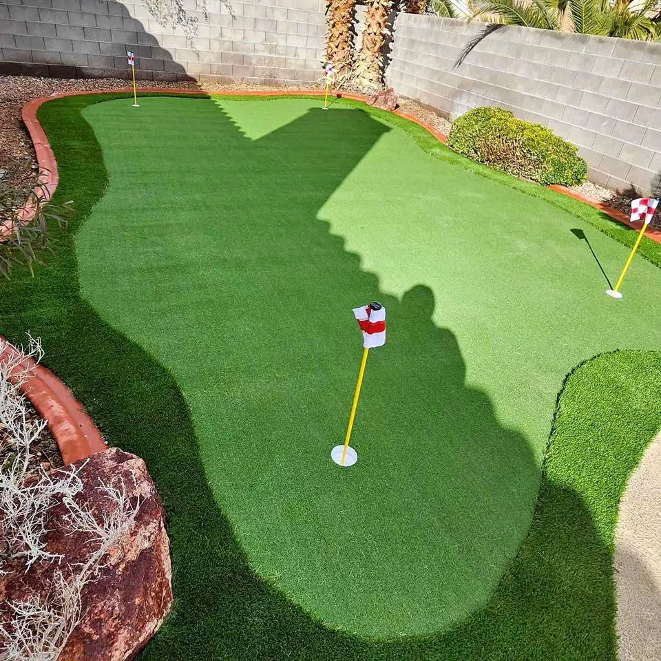 A backyard putting green with artificial turf, three golf holes marked by red and white checkered flags, bordered by a gray cinder block wall and landscaping with rocks and plants—perfect for golf enthusiasts in Mesa AZ.