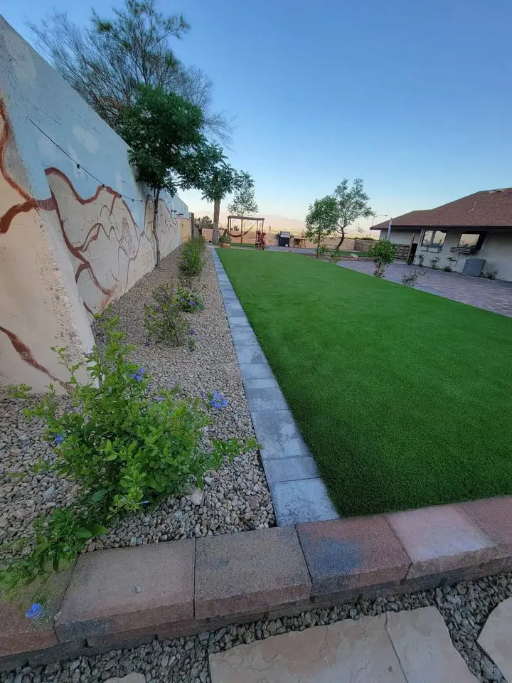 A backyard in Mesa AZ features a lush green lawn with artificial turf bordered by a stone walkway, a colorful garden bed, decorative wall art, and a charming house with a tiled roof in the background.