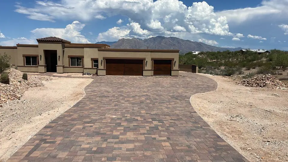 Single-story modern house with a three-car garage and tan exterior sits at the end of a wide, curved paved driveway in Gilbert AZ, surrounded by desert landscaping, turf options, and distant mountains under a partly cloudy sky.