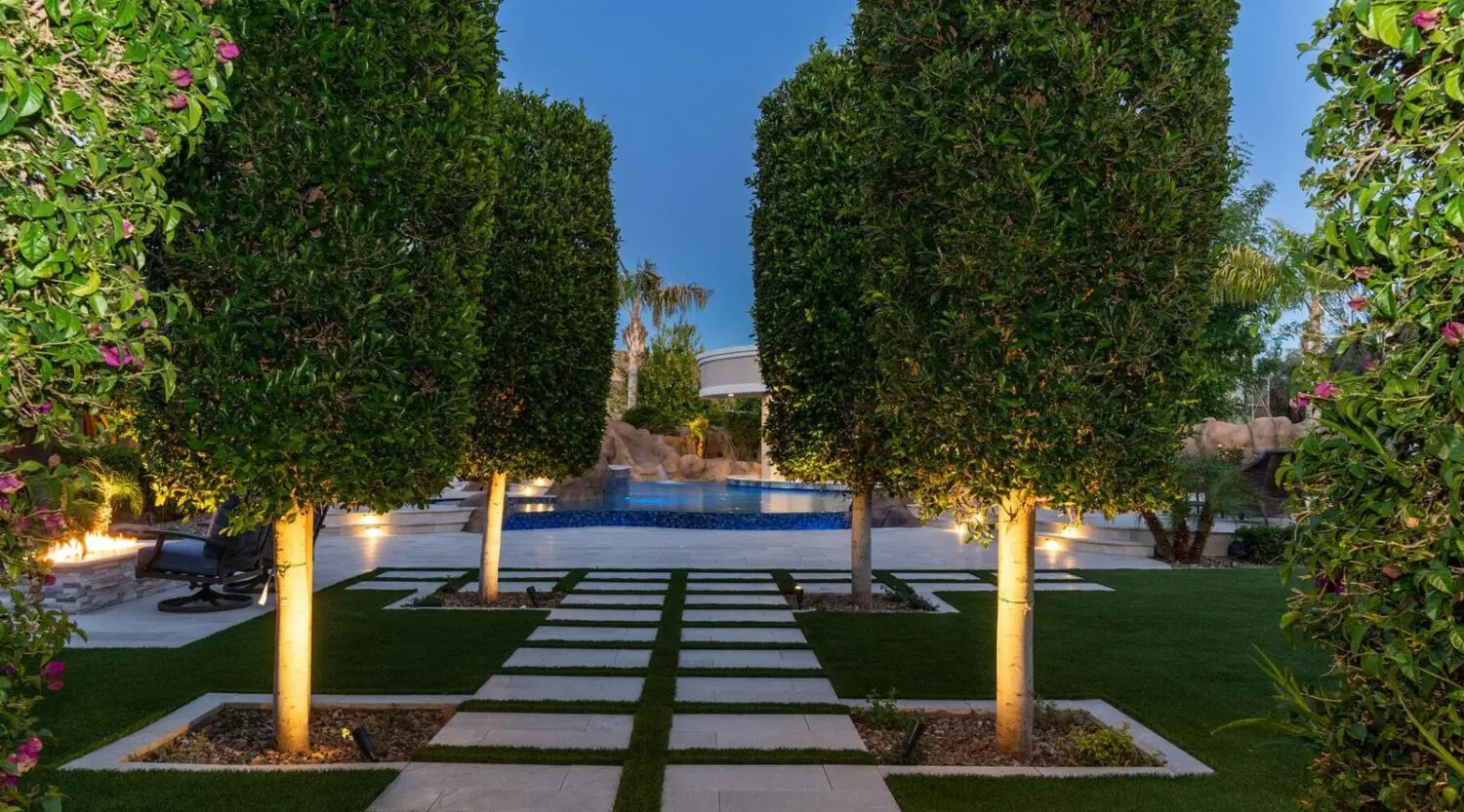 A landscaped backyard in Gilbert AZ features tall, neatly trimmed trees lining square stone pavers leading to a lit swimming pool, surrounded by greenery and lush turf options at dusk.