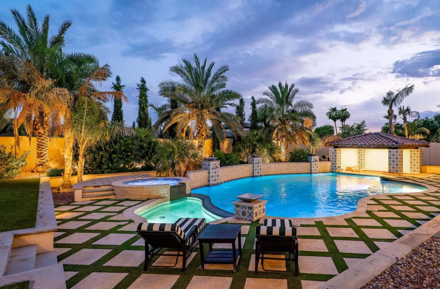 Luxurious backyard in Gilbert AZ with a large, illuminated pool and hot tub, surrounded by palm trees and lush landscaping. Enjoy turf options from an expert artificial grass installer beside two striped lounge chairs on a stone patio at dusk.
