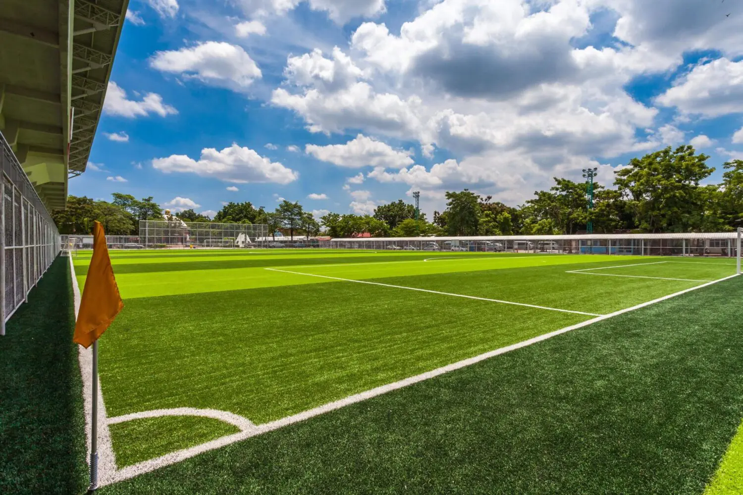 A brightly lit outdoor soccer field with green artificial turf showcases quality turf options. A yellow corner flag, white boundary lines, and a blue sky with scattered clouds complete the scene in Gilbert AZ. Trees and a fence surround the field.