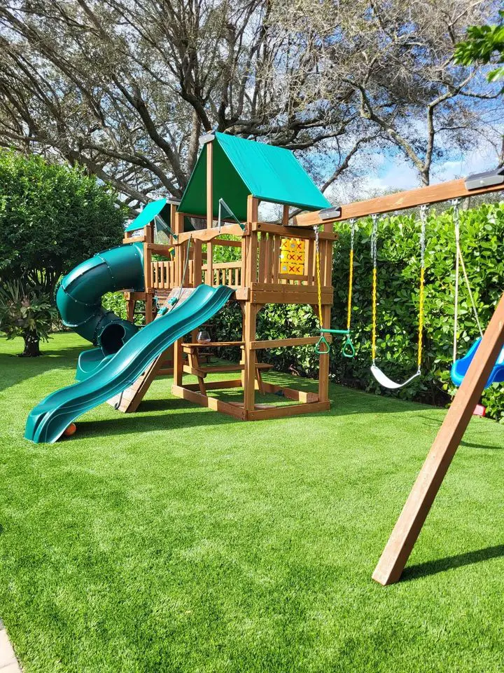 A wooden backyard playset with a green slide, spiral tube slide, swings, ladders, and a small clubhouse sits on vibrant artificial grass. Consider local turf options from an experienced artificial grass installer in Gilbert AZ.