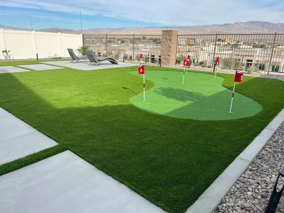 A backyard in Gilbert AZ with a small artificial putting green featuring four red golf flags, surrounded by lush turf options, lounge chairs, and a paved patio area, with a fence and scenic mountains in the background.