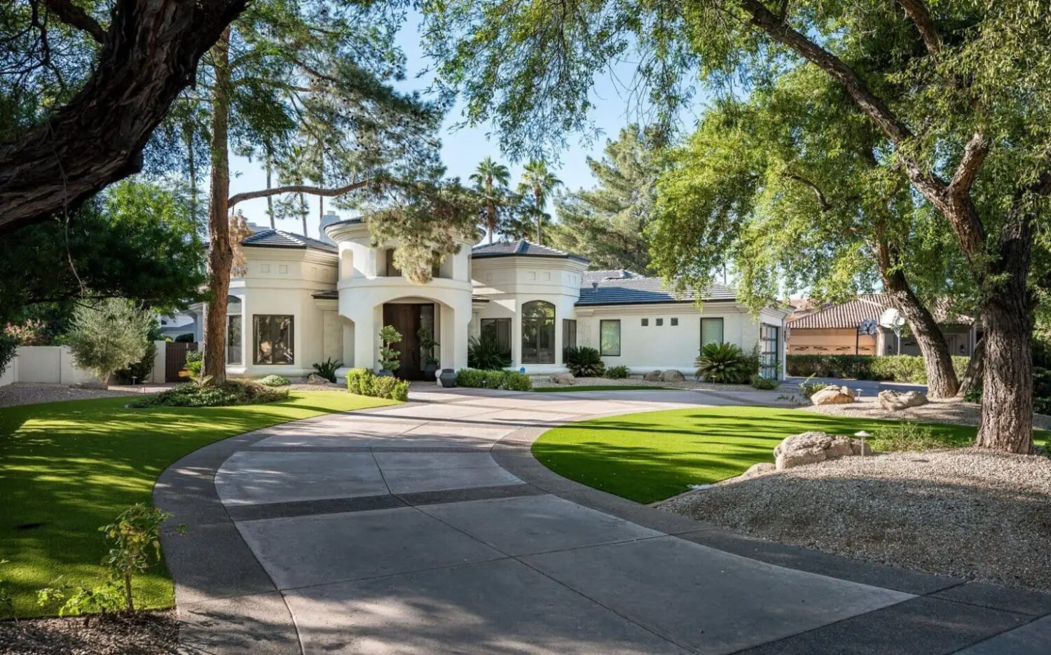 A large, modern white house with tall windows and a circular entryway sits behind a sweeping curved driveway in San Tan Valley, surrounded by manicured lawns, artificial grass, rocks, and tall green trees under a clear sky.