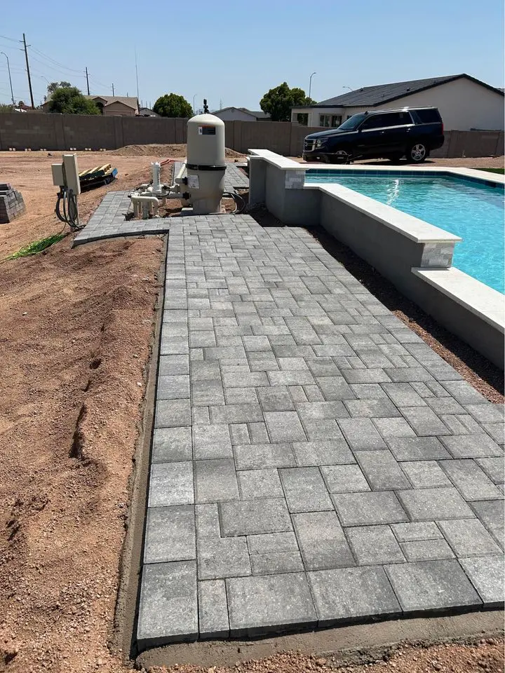 A backyard under construction in Queen Creek AZ features a newly installed gray stone paver walkway beside a swimming pool, with unfinished landscaping and loose dirt. A black SUV and houses are visible in the background, highlighting recent paver installation work.