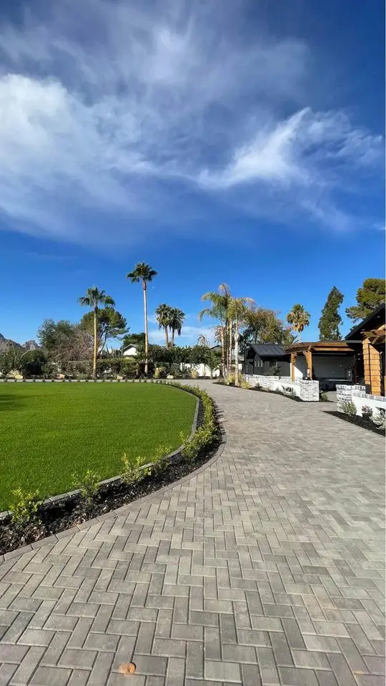 A paved walkway curves past green grass and palm trees under a bright blue sky with scattered clouds, beside a building with white and wooden walls, showcasing expert paver installation in Queen Creek AZ.