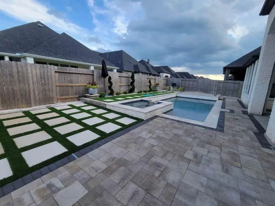 Modern backyard in Queen Creek AZ with a small pool, attached hot tub, paver patio with geometric paver installation and artificial grass, wooden fence, sculptural planters, and neighboring houses under a cloudy sky.