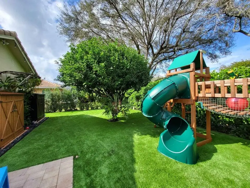 A backyard in Chandler AZ with lush artificial grass, a large tree, and a wooden playset featuring a green spiral slide and climbing area. Bushes border the yard, and a wooden fence runs along the left side—perfect turf solutions for family fun.