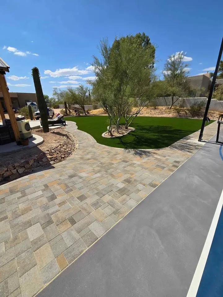 A backyard in Queen Creek AZ features a stone paver patio, artificial turf lawn, desert landscaping with cacti and trees, and a partial view of a concrete surface under a clear blue sky. Perfect for outdoor gatherings and relaxing evenings.
