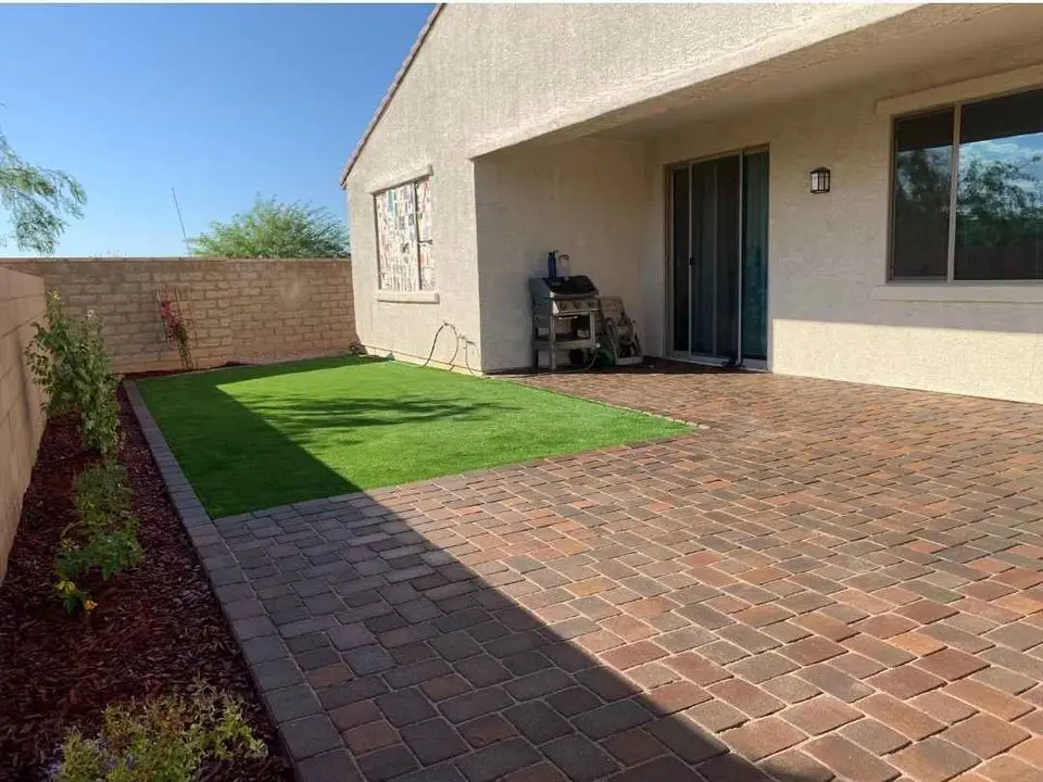 A backyard in Queen Creek AZ featuring a paver patio, a small section of artificial grass, a grill, plants along the edge, and a beige house with sliding glass doors and a window.